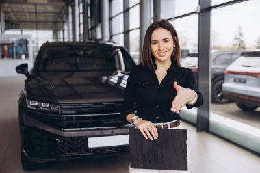 Satıcı kadın, araba galerisinde yeni bir aracın önünde klasörü tutarken müşteriye el sıkışma teklif ediyor.