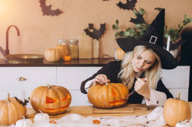 Cadı şapkası takan bir kadın neşeli bir mutfak ortamında Cadılar Bayramı için balkabağı yüzünü oyuyor.