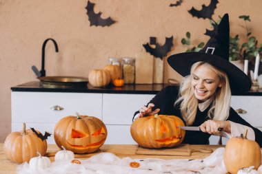 Cadı şapkası takan genç bir kadın Cadılar Bayramı partisi için balkabağı oyuyor.