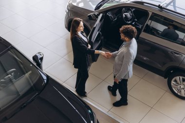 Modern bir araba galerisinde satış elemanı ve müşteri el sıkışıyor. Yüksek açılı bir bakış açısıyla anlaşmanın başarıyla sonuçlanmasını kutluyorlar.