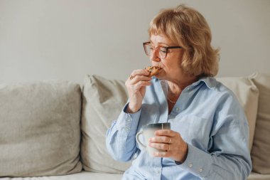 Yaşlı bir kadın rahat bir kanepede dinleniyor, kurabiye ve sıcak bir içeceğin tadını çıkarıyor, evde sakin bir anın tadını çıkarıyor.
