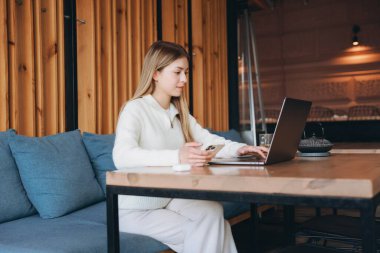 Rahat bir kafede uzaktan çalışan serbest çalışan, rahat bir atmosfer ve verimli bir ortamın keyfini sürerken dizüstü bilgisayar ve akıllı telefon kullanan serbest çalışan biri.