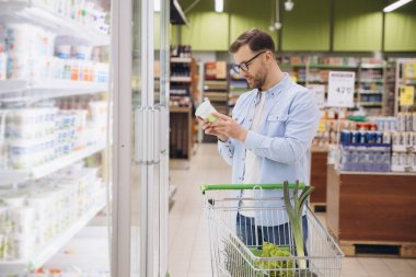 Man choosing a yogurt in refrigerated section of supermarket, reading label and pushing shopping cart