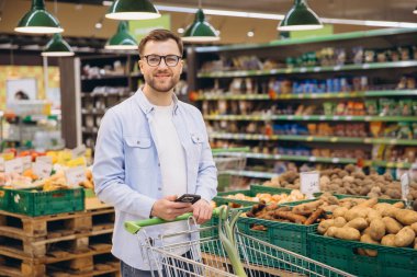 Man using smartphone and pushing shopping cart while shopping for groceries in supermarket