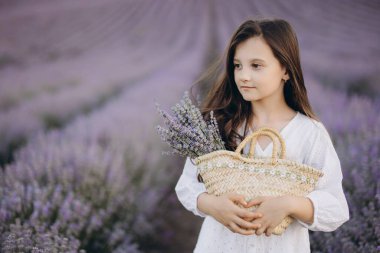 Girl in a flowing white dress, holding a straw bag and a vibrant lavender bouquet, enjoying the beauty of a blooming lavender field
