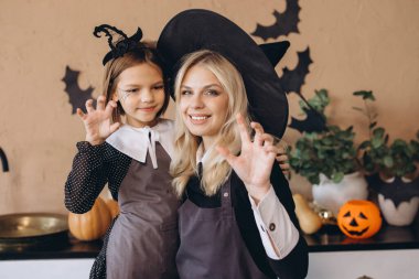 Mom and daughter making scary faces and hand gestures, wearing witch costumes and preparing for a Halloween party at home