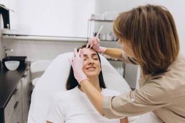 Beautician drawing marks on forehead of young woman before cosmetic injections in beauty salon