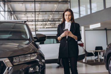 Saleswoman using mobile phone in a car dealership, standing next to a new car, ready to assist customers