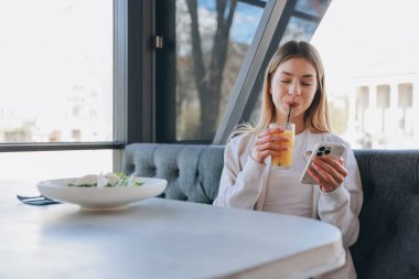 Businesswoman drinking orange juice and using mobile phone in a restaurant, enjoying a healthy lunch break