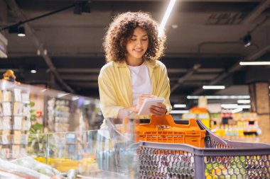 Smiling curly woman using digital tablet checking shopping list while pushing trolley in supermarket