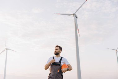 Engineer holding a helmet and gazing upward at towering wind turbines in a wind farm, highlighting the promise of renewable energy