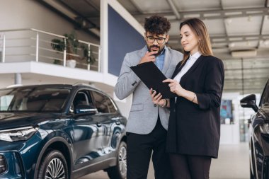Salesperson explaining car features to a thoughtful client holding a clipboard, engaging in a detailed discussion inside a modern dealership
