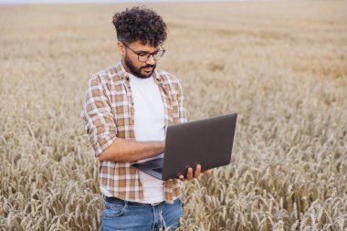 Young Arab agronomist working with laptop in wheat field, implementing modern technology in agriculture for efficient crop growth and sustainable practices