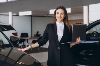 Smiling saleswoman holding a laptop while presenting a new car to potential clients in a modern dealership showroom