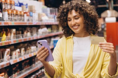 Young woman using a smartphone and credit card while shopping for groceries in a vibrant supermarket, enjoying a modern retail experience