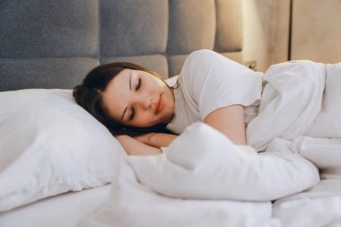 Young woman resting in comfortable bed with white bedding enjoying good night sleep in bedroom