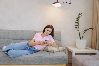 Smiling woman lying comfortably on sofa using smartphone, enjoying leisure time in cozy home environment