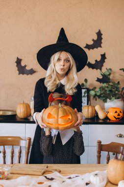 Mother wearing witch costume and daughter with devil horns holding jack-o'-lantern in decorated kitchen for Halloween