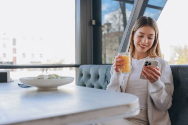 Happy woman savoring breakfast in a stylish restaurant, enjoying fresh orange juice while browsing her smartphone and connecting online