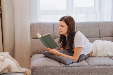Young woman enjoying a relaxing afternoon reading a book while lying comfortably on the sofa in her modern living room