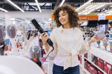 Smiling curly haired woman comparing hair dryer brush and hair curler, shopping for hair styling products in consumer electronics store