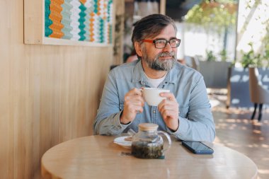 Bearded senior man savoring a cup of tea in a modern cafe, embracing a peaceful moment of relaxation and quiet contemplation
