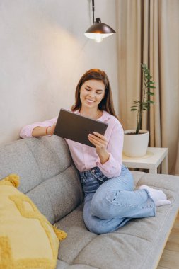 Young woman relaxing on comfortable sofa using digital tablet, enjoying online entertainment or working remotely from home