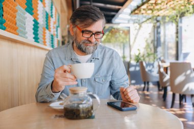 Bearded senior man savoring a cup of tea while browsing his smartphone in a modern cafe, enjoying a relaxed moment in a cozy atmosphere