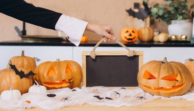 Woman holding an empty blackboard surrounded by carved pumpkins and festive Halloween decorations on a kitchen table, creating a spooky atmosphere