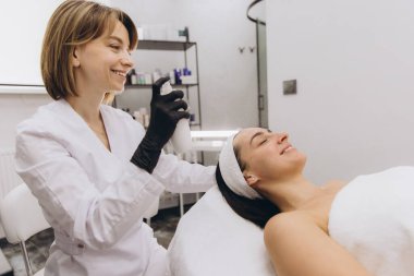 Smiling beautician performing skincare treatment on a client lying on a massage table in a professional beauty salon