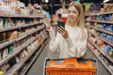 Markette alışveriş yapan genç bir kadın, akıllı telefon ve kredi kartıyla koridorlarda gezinirken bakkaliye alışverişi yapıyor.