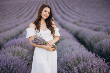 Young woman wearing a flowing white dress, holding a fragrant lavender bouquet while surrounded by vibrant lavender blooms in a scenic field