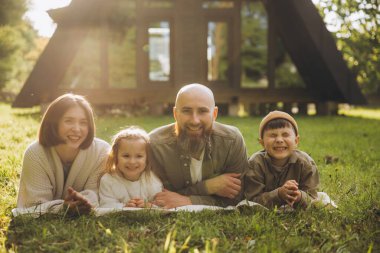 Smiling family relaxing on the grass in front of their cozy cabin, enjoying a sunny vacation day filled with laughter and joy