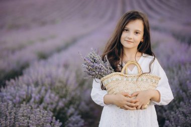 Girl wearing a flowing white dress, holding a wicker bag and a vibrant lavender bouquet while wandering through a picturesque lavender field