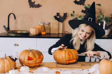 Young woman wearing a witch hat, happily carving a pumpkin for Halloween party decorations in her cozy kitchen at home