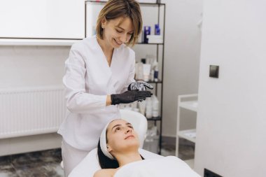 Smiling cosmetologist wearing black gloves is preparing a cleansing facial treatment for her customer lying on a massage table in a beauty salon