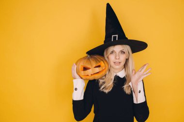 Halloween witch wearing black hat and dress holding carved pumpkin with surprised expression on yellow background