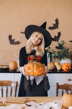 Mother wearing witch costume and daughter with devil horns holding carved pumpkin with candle inside during Halloween preparation