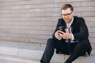 Businessman with loose tie and eyeglasses using mobile phone app while sitting on curb near gray brick wall
