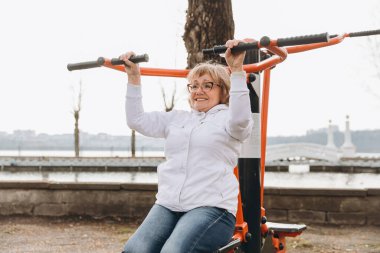 Elderly woman exercising on outdoor gym equipment by the river, promoting healthy lifestyle and active aging