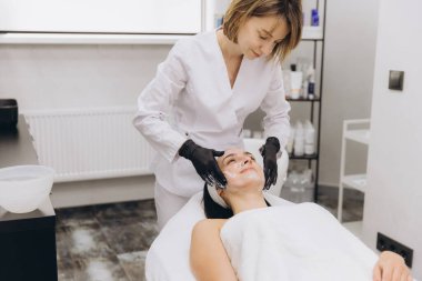 Beautician wearing black gloves applying a face mask to a young woman lying on a treatment table in a professional beauty salon