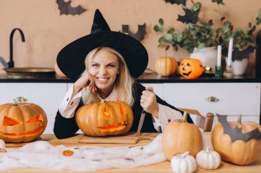 Young woman wearing witch costume carving pumpkins for Halloween party, holding knife and looking mischievous