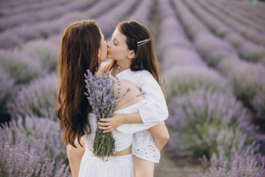 Mother carrying her daughter on her back, kissing her in a lavender field while holding a bouquet