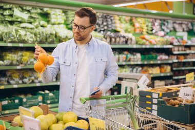 Customer selecting ripe oranges in a grocery store, checking a shopping list while pushing a cart filled with fresh produce