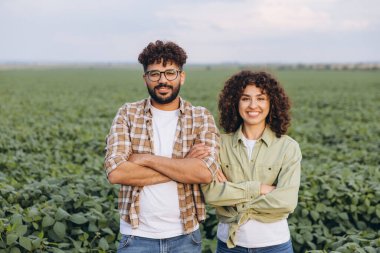 Interracial team of two agronomists standing confidently with arms crossed, posing in a lush, cultivated soybean field under a clear summer sky