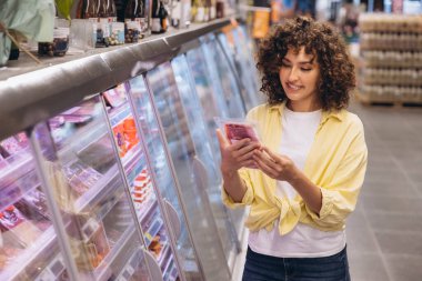 Smiling woman with curly hair selecting packaged meat from the refrigerated section while shopping for groceries in a supermarket aisle