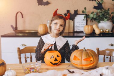 Girl wearing devil horns headband and halloween costume preparing a pumpkin basket with candy, sitting at a wooden table with carved pumpkins