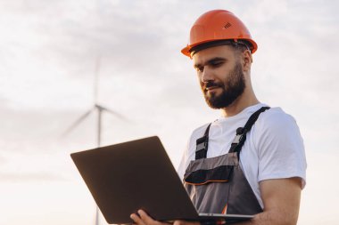 Engineer working on a laptop while overseeing a wind turbine in the background, contributing to sustainable energy solutions and innovation