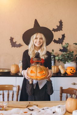 Mother wearing witch hat and daughter with devil horns holding carved pumpkin with illuminated face during Halloween celebration in decorated kitchen