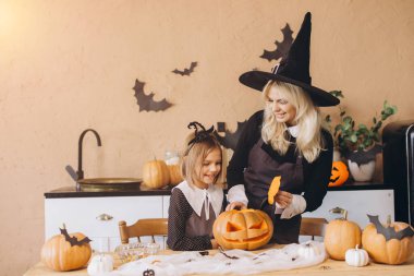 Mother in a witch costume guiding her daughter while carving a pumpkin for a festive Halloween party, creating joyful family memories together